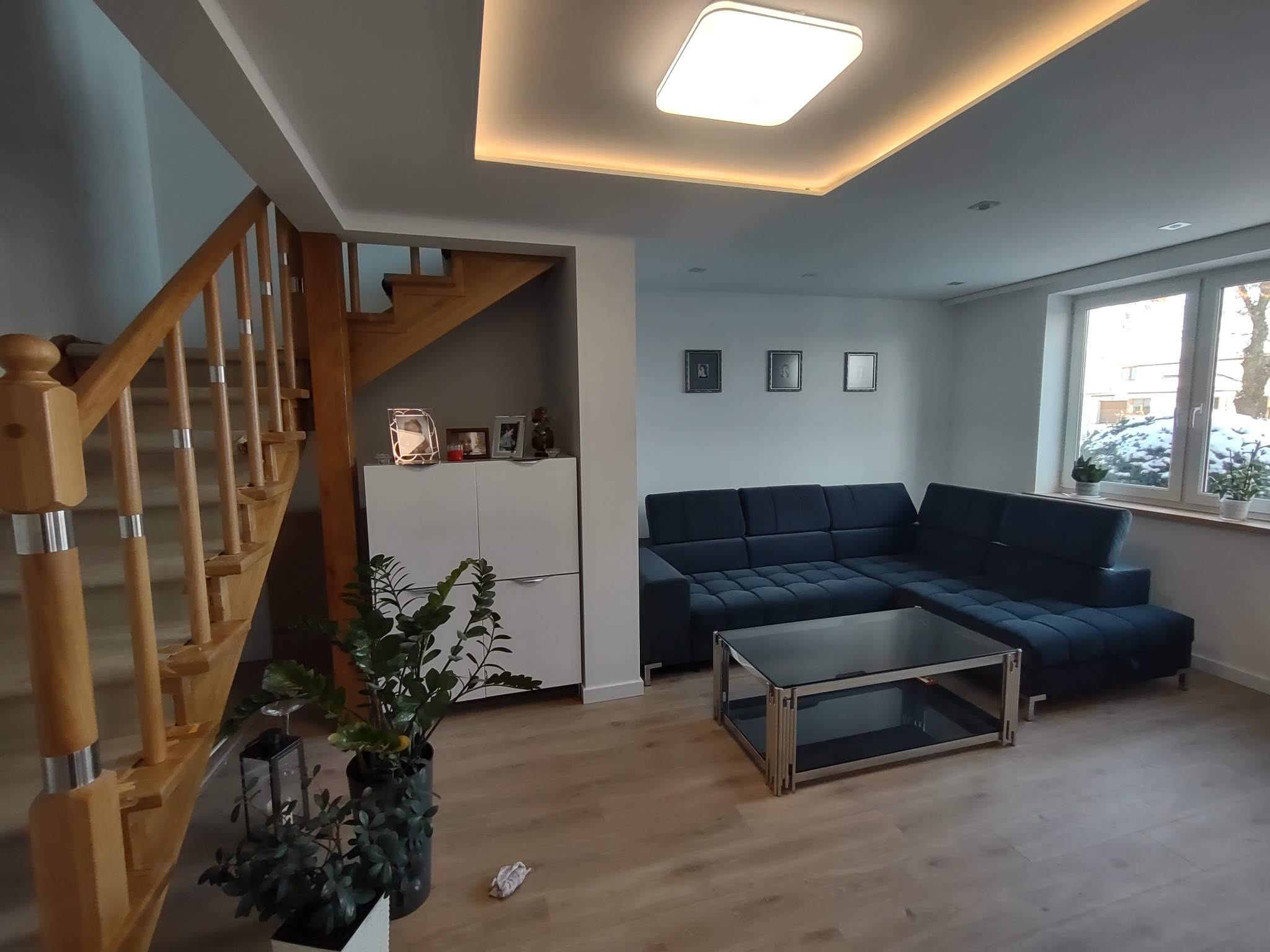 Nowoczesny salon z drewnianymi schodami, narożną sofą w odcieniach granatu i szklanym stolikiem kawowym, oświetlony plafonem z ciepłym światłem LED.