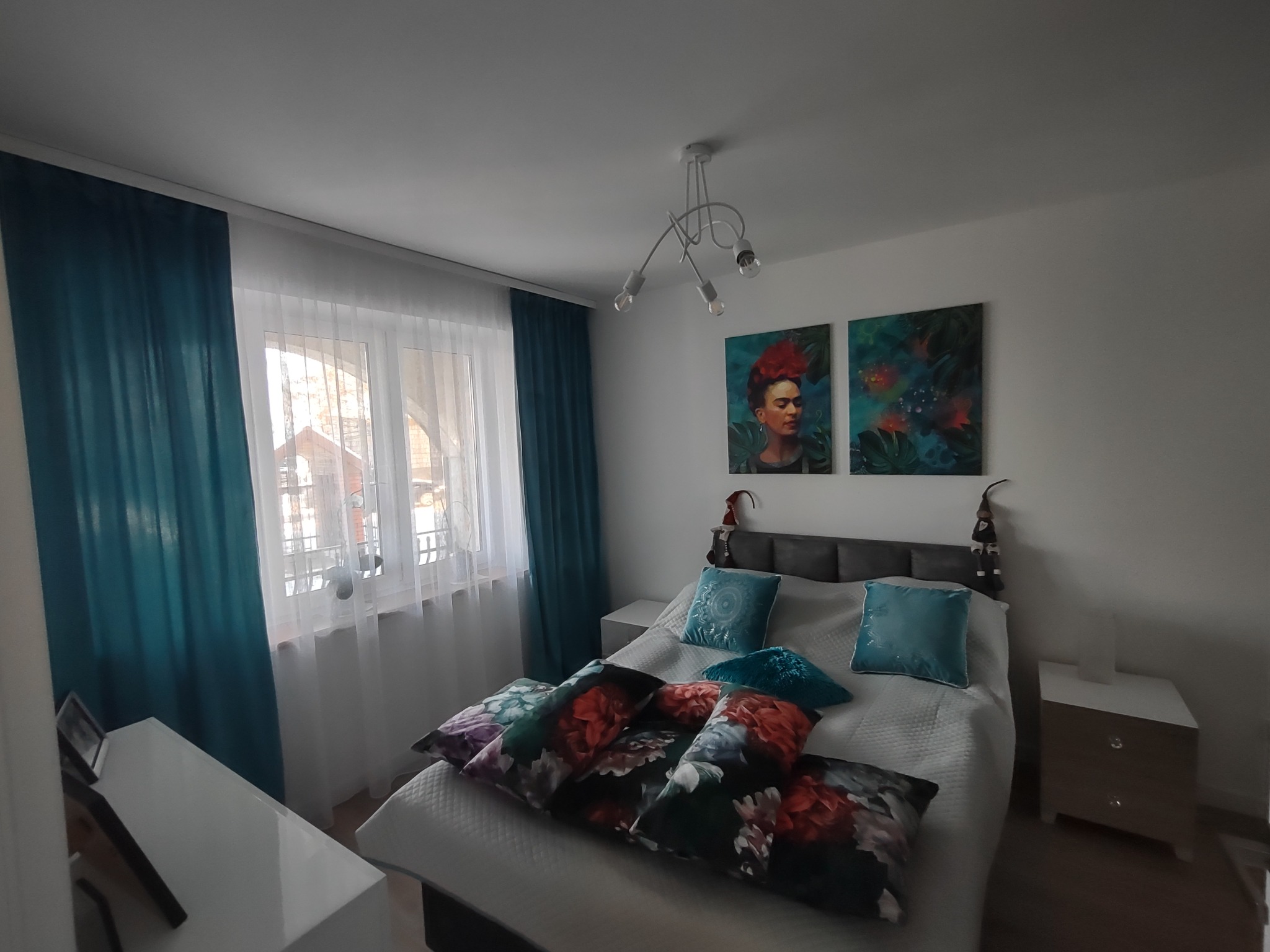 Jasna sypialnia z łóżkiem z szarą ramą, turkusowymi zasłonami i poduszkami oraz dwoma obrazami z motywem Fridy Kahlo nad wezgłowiem.