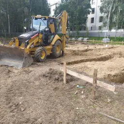 Żółta koparko-ładowarka CAT 432E na placu budowy z wykopami pod fundamenty, w tle budynek mieszkalny i drzewa.