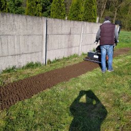 Kena - Mężczyzna w kamizelce i jeansach używa glebogryzarki spalinowej do przygotowania gruntu wzdłuż betonowego ogrodzenia, widoczny cień osoby robiącej zdjęcie.