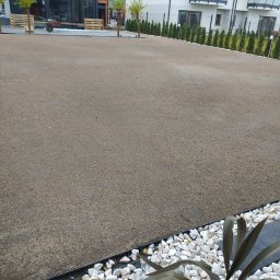 Kena - Przygotowanie podłoża pod trawnik: wyrównana powierzchnia z piasku i humusu, obrzeża z białych kamieni i zielonych tuj, w tle nowoczesny dom.