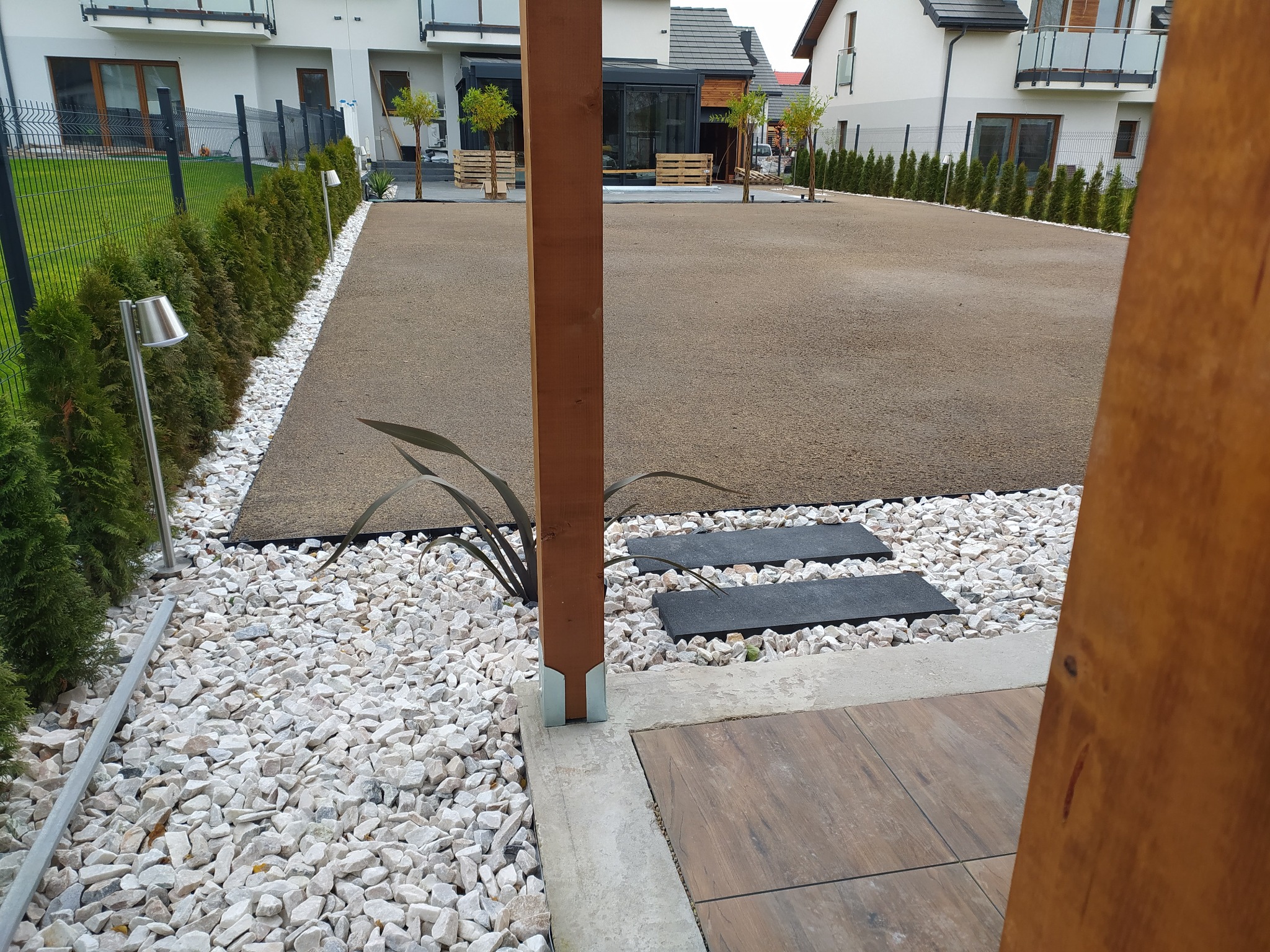 Wykończony ogród z równą, brązową powierzchnią, obrzeżami z białych kamieni, ścieżką z czarnych płyt i drewnianą pergolą w tle nowoczesnych domów.