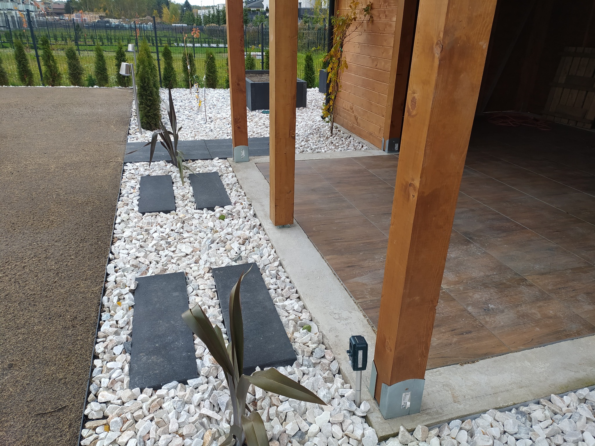 Nowoczesna aranżacja ogrodu z drewnianą altaną, ścieżką z ciemnych płyt ułożonych na białym żwirze i ozdobnymi roślinami.