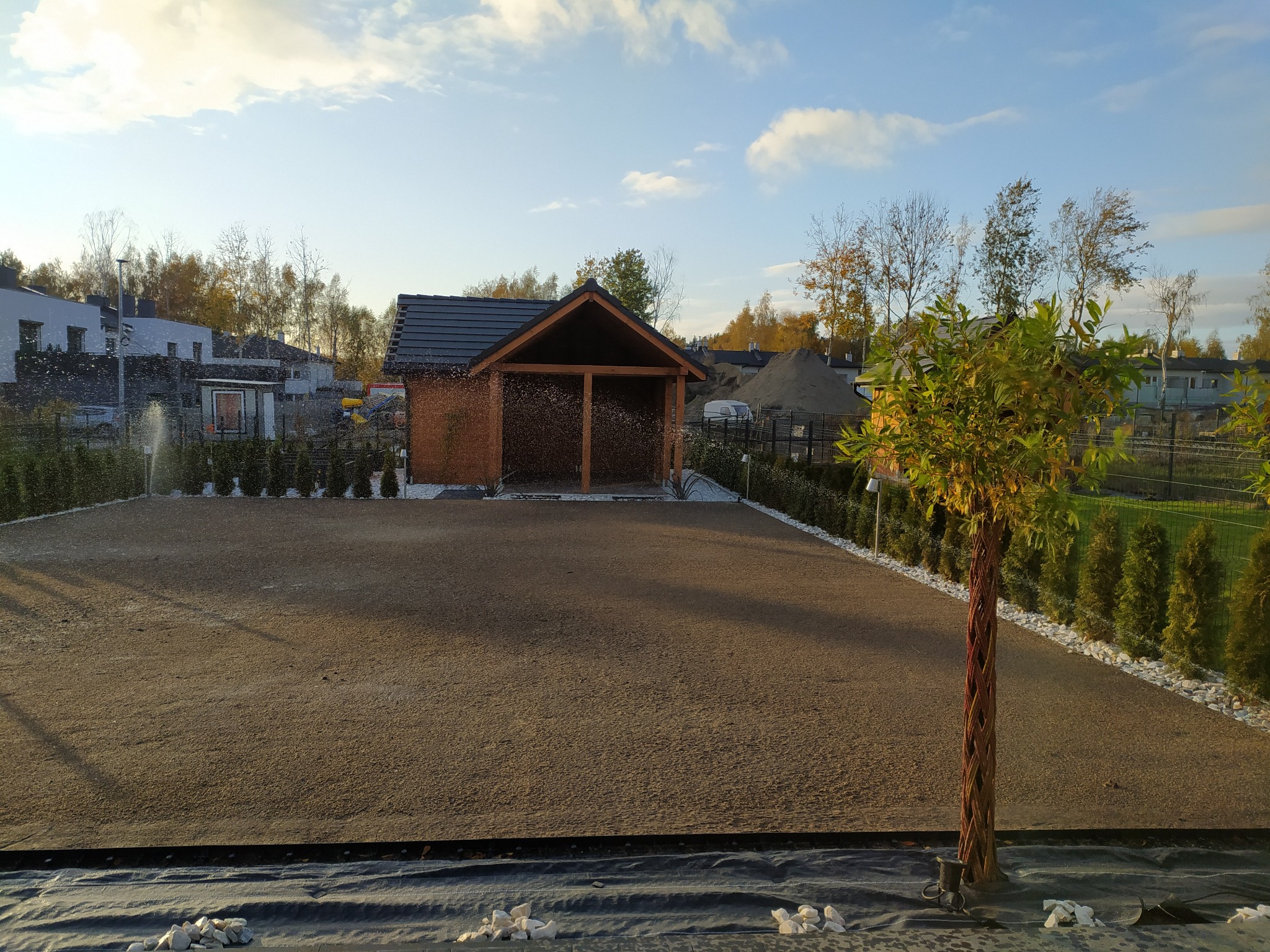 Przygotowany teren pod trawnik z widoczną drewnianą altaną, nawadnianiem i ozdobnym drzewem na pierwszym planie.