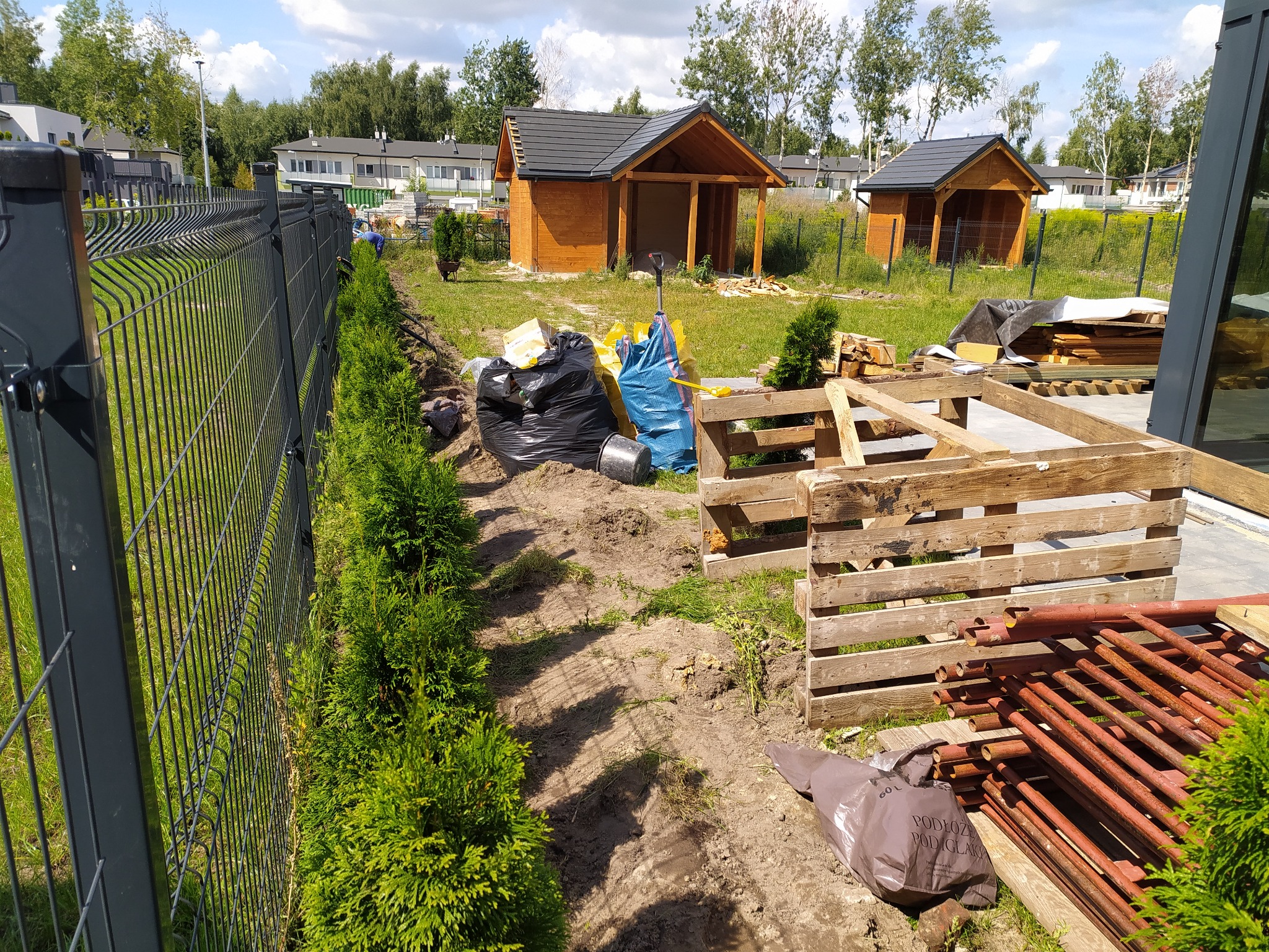 Przygotowanie terenu pod nasadzenia w ogrodzie: widoczny wykop wzdłuż metalowego ogrodzenia, posadzone małe tuje, ziemia, worki z ziemią, drewniane konstrukcje i dwa drewniane domki narzędziowe w tle.