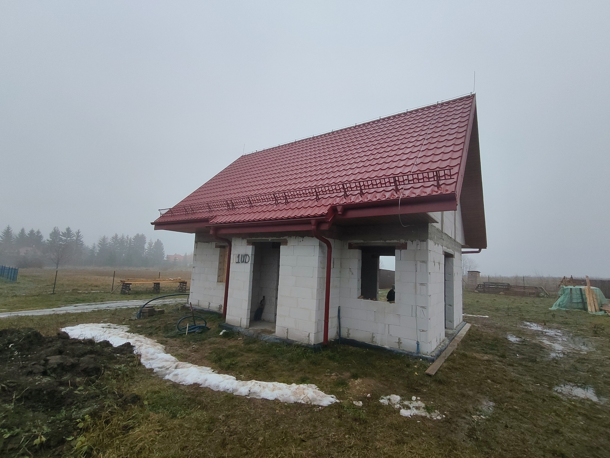 Niedokończony dom z czerwonym dachem z blachodachówki, w trakcie budowy, z widocznymi bloczkami i oknami bez szyb, na zewnątrz widać błoto i resztki śniegu.