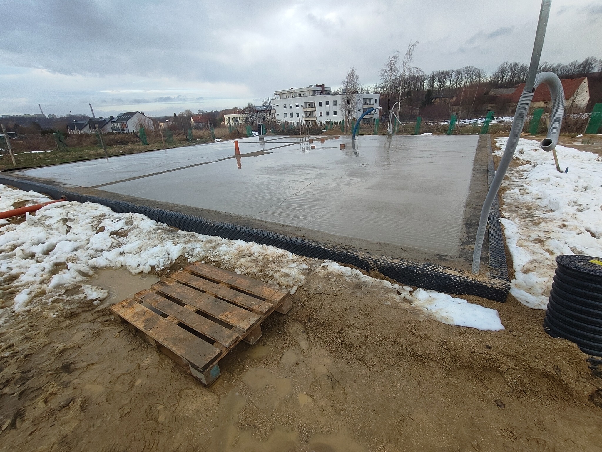 Wylana płyta fundamentowa pod budowę domu jednorodzinnego, widoczne zbrojenia i elementy instalacji. Na pierwszym planie paleta budowlana i resztki śniegu.
