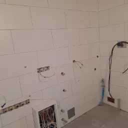Ściany wyłożone białymi, prostokątnymi płytkami w trakcie prac wykończeniowych; widoczne wystające przewody elektryczne, rury instalacji wodnej oraz częściowo zabudowana skrzynka z instalacją.