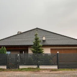 Nowoczesne, poziome przęsła ogrodzeniowe w antracytowym kolorze przed domem z dwuspadowym dachem krytym dachówką ceramiczną; widoczna furtka z domofonem.
