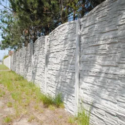 Długi, szary mur z betonowych element&oacute;w imitujących kamień, oddzielający posesję od ulicy, z widocznymi drzewami w tle.