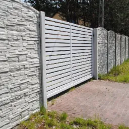 Nowoczesna, biała brama przesuwna z poziomych profili, zamontowana między betonowymi słupkami i segmentami ogrodzenia z imitacją kamienia łupanego, na podjeździe z kostki brukowej.