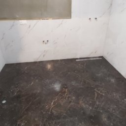 M.C.Remont Michał Ciszek - Wnętrze łazienki w trakcie wykańczania, z białymi płytkami imitującymi marmur na ścianach i ciemną, marmurkową podłogą, widoczne wystające rury instalacji wodnej i fragment listwy odpływowej...