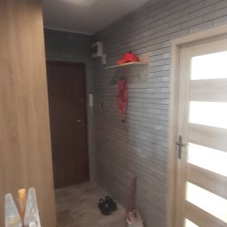 M.C.Remont Michał Ciszek - Korytarz w trakcie remontu z drabiną malarską, wiadrem z farbą i nową okładziną ścienną imitującą cegłę oraz płytkami podłogowymi w odcieniach beżu.