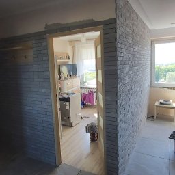 M.C.Remont Michał Ciszek - Korytarz w trakcie remontu z fragmentami ściany wyłożonymi szarą cegłą dekoracyjną, widoczne otwarte drzwi do pokoju i łazienki, oraz okno z widokiem na zieleń.