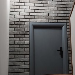M.C.Remont Michał Ciszek - Szara ściana z cegły dekoracyjnej, szare drzwi wewnętrzne z czarną klamką i fragment drewnianej podłogi, oświetlenie punktowe na ścianie