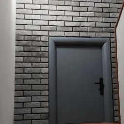 Szara ściana z cegły dekoracyjnej, szare drzwi wewnętrzne z czarną klamką i fragment drewnianej podłogi, oświetlenie punktowe na ścianie