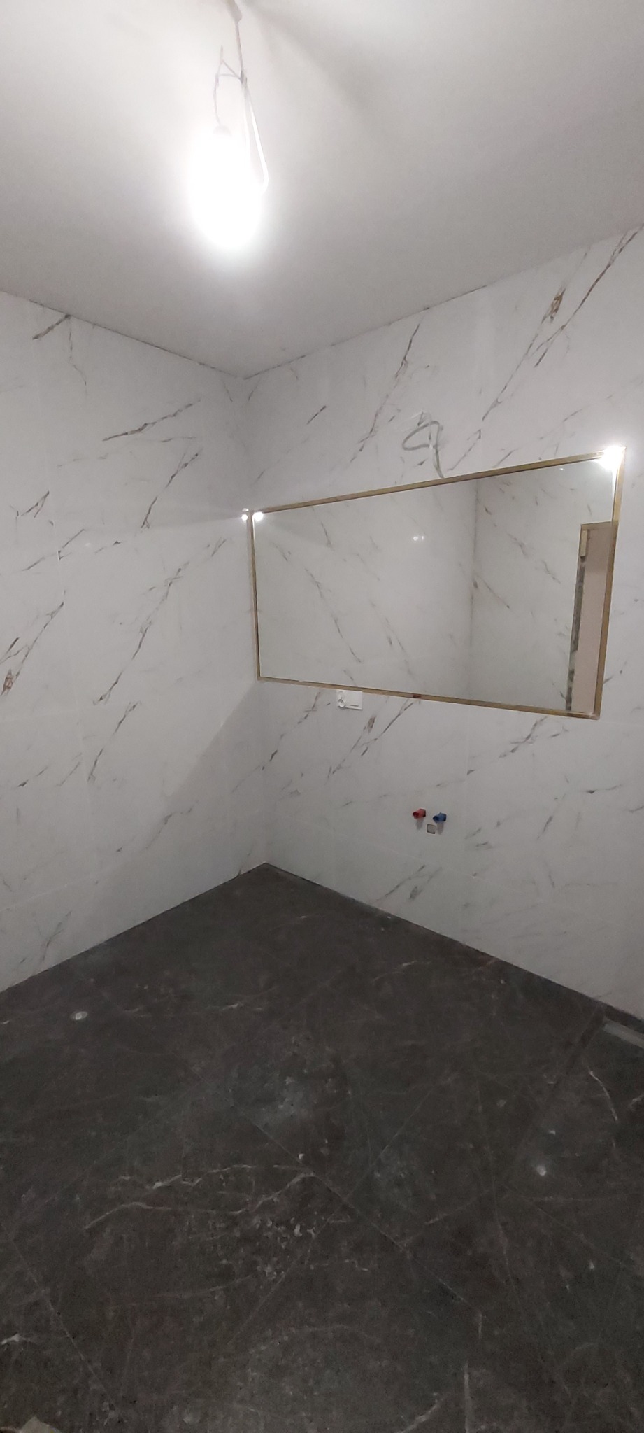 Wykończona łazienka z marmurowymi płytkami ściennymi i podłogowymi w odcieniach bieli i szarości, z zamontowanym lustrem w złotej ramie i wystającymi rurami instalacji wodnej.