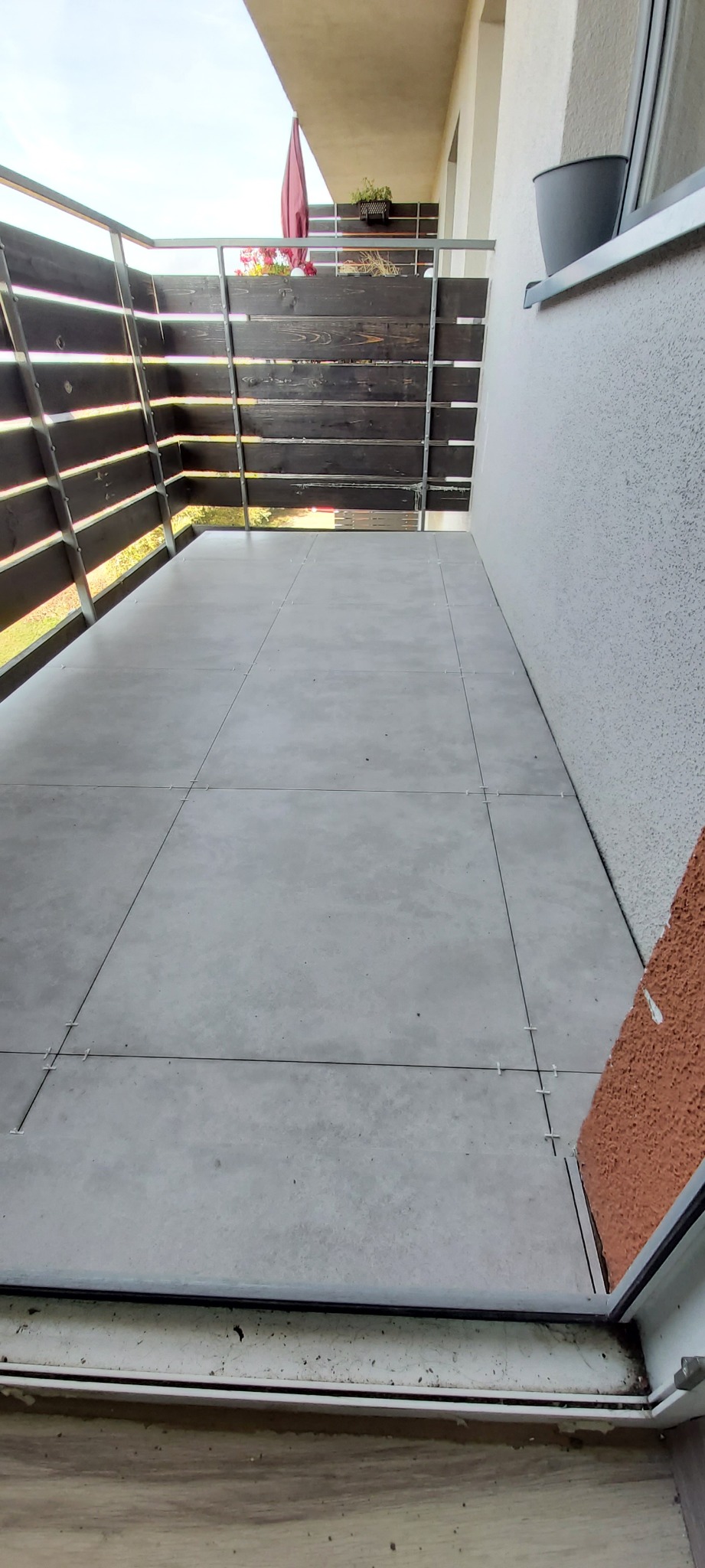 Układanie szarych płytek na balkonie w Inowrocławiu z użyciem krzyżyków dystansowych, widok na drewnianą balustradę i doniczkę z kwiatami.