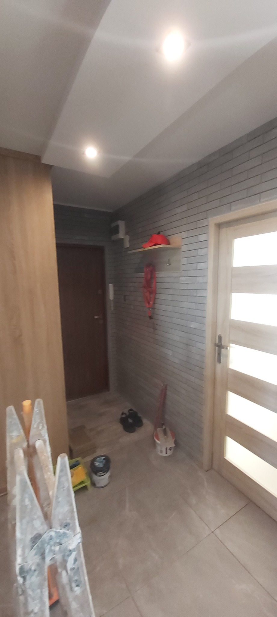 Korytarz w trakcie remontu z drabiną malarską, wiadrem z farbą i nową okładziną ścienną imitującą cegłę oraz płytkami podłogowymi w odcieniach beżu.