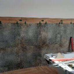 Ściana w trakcie wykańczania płytkami z dekoracyjnym wzorem, użyte zielone klipsy poziomujące, widoczny poziomica i opakowania płytek.