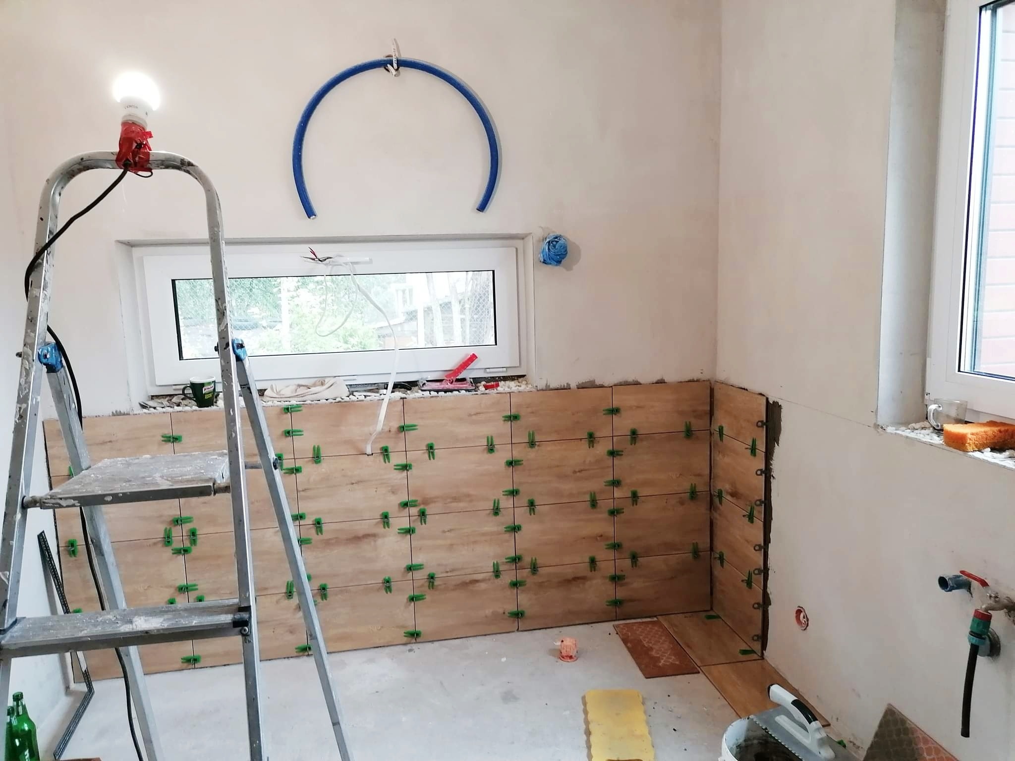Proces układania płytek imitujących drewno na ścianie w łazience, widoczne krzyżyki dystansowe, drabina z prowizorycznym oświetleniem i niebieski wąż na ścianie.