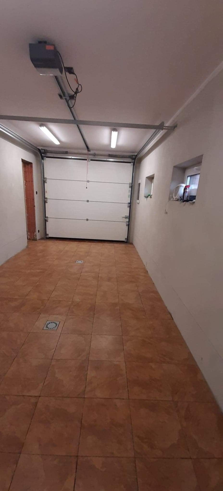 Wnętrze garażu z brązowymi płytkami podłogowymi, białą segmentową bramą garażową i automatyką pod sufitem. Ściany pomalowane na biało, widoczne okna i drzwi w surowym stanie.