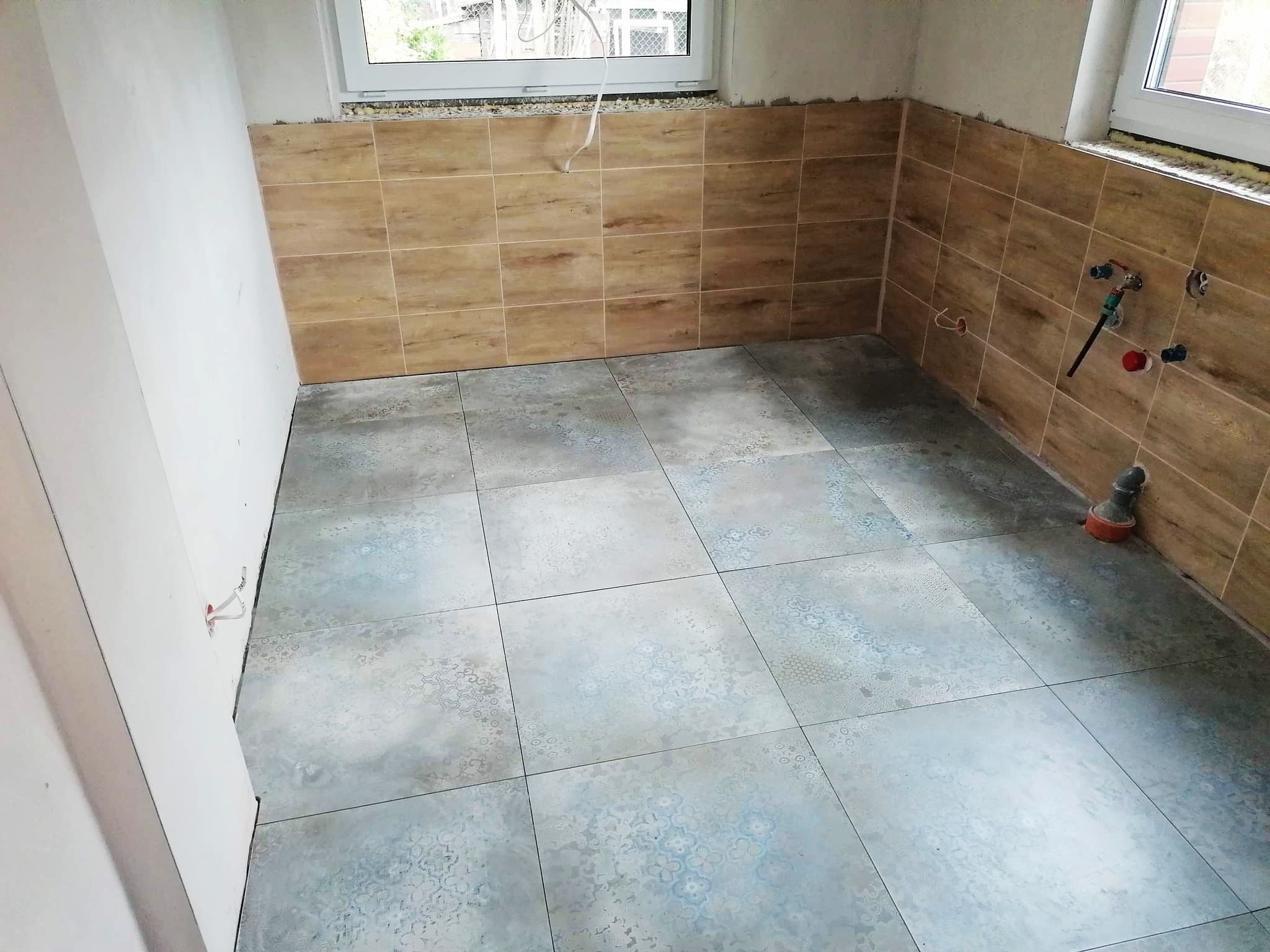 Świeżo położone, wzorzyste płytki podłogowe w pomieszczeniu z brązowymi płytkami ściennymi imitującymi drewno i widocznymi instalacjami hydraulicznymi.