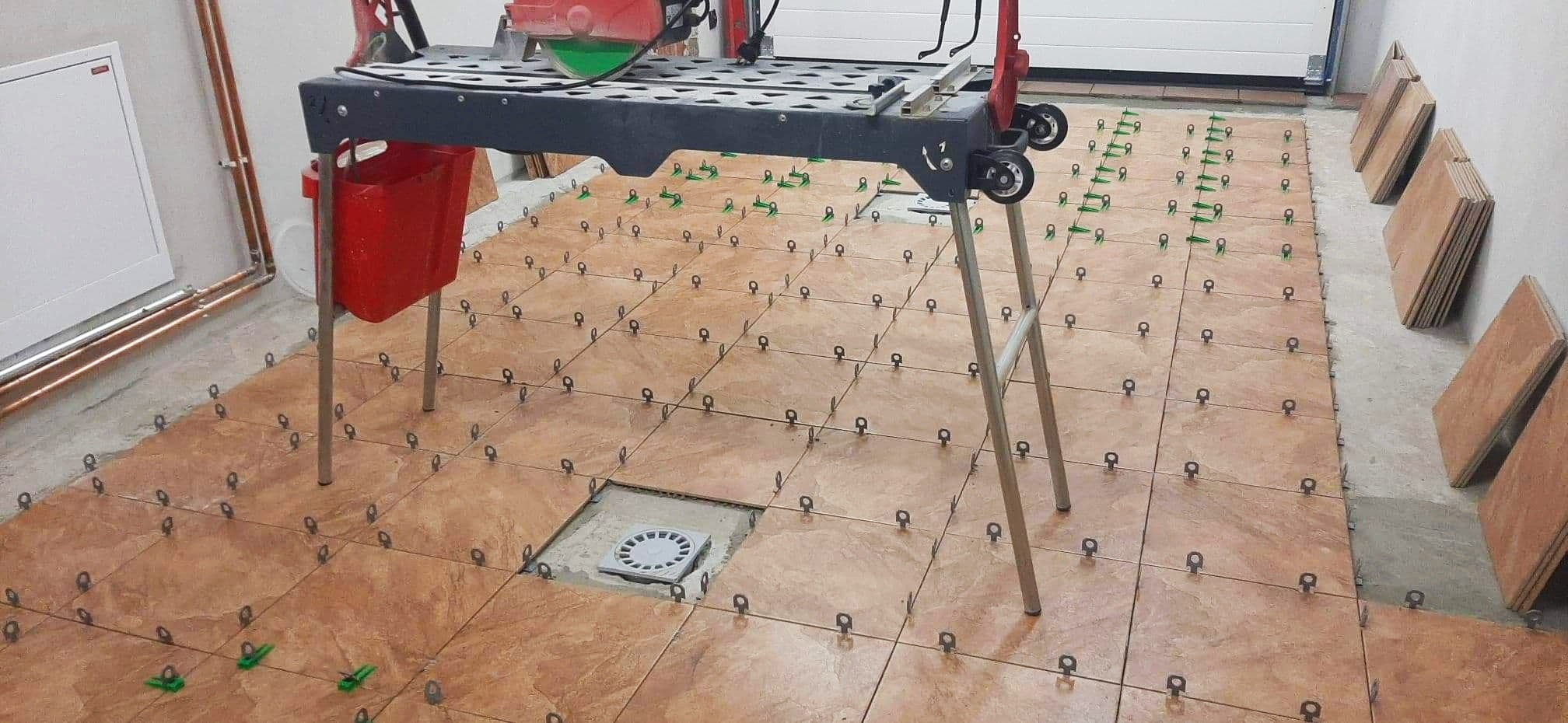 Brązowe płytki podłogowe układane w garażu z wykorzystaniem systemu poziomowania, widoczna maszyna do cięcia płytek i odpływ liniowy w podłodze.