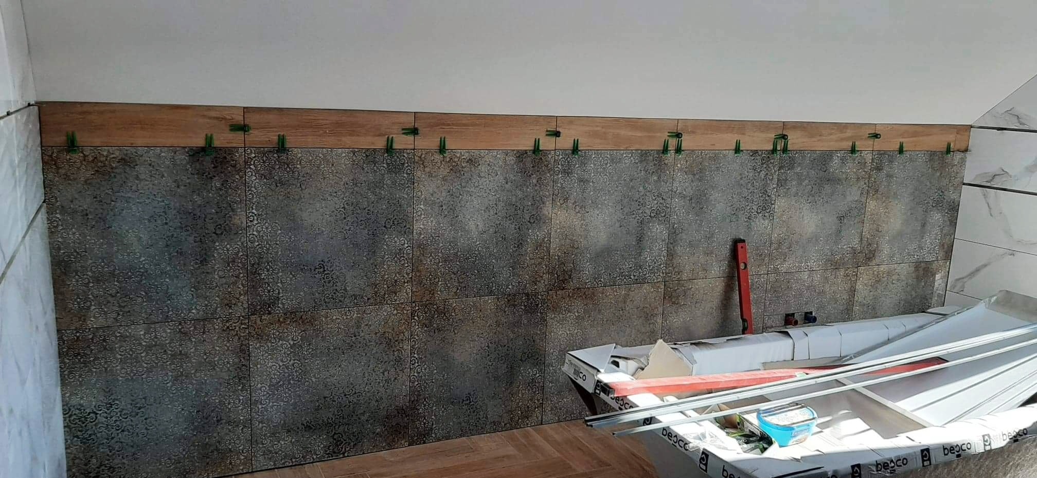 Ściana w trakcie wykańczania płytkami z dekoracyjnym wzorem, użyte zielone klipsy poziomujące, widoczny poziomica i opakowania płytek.