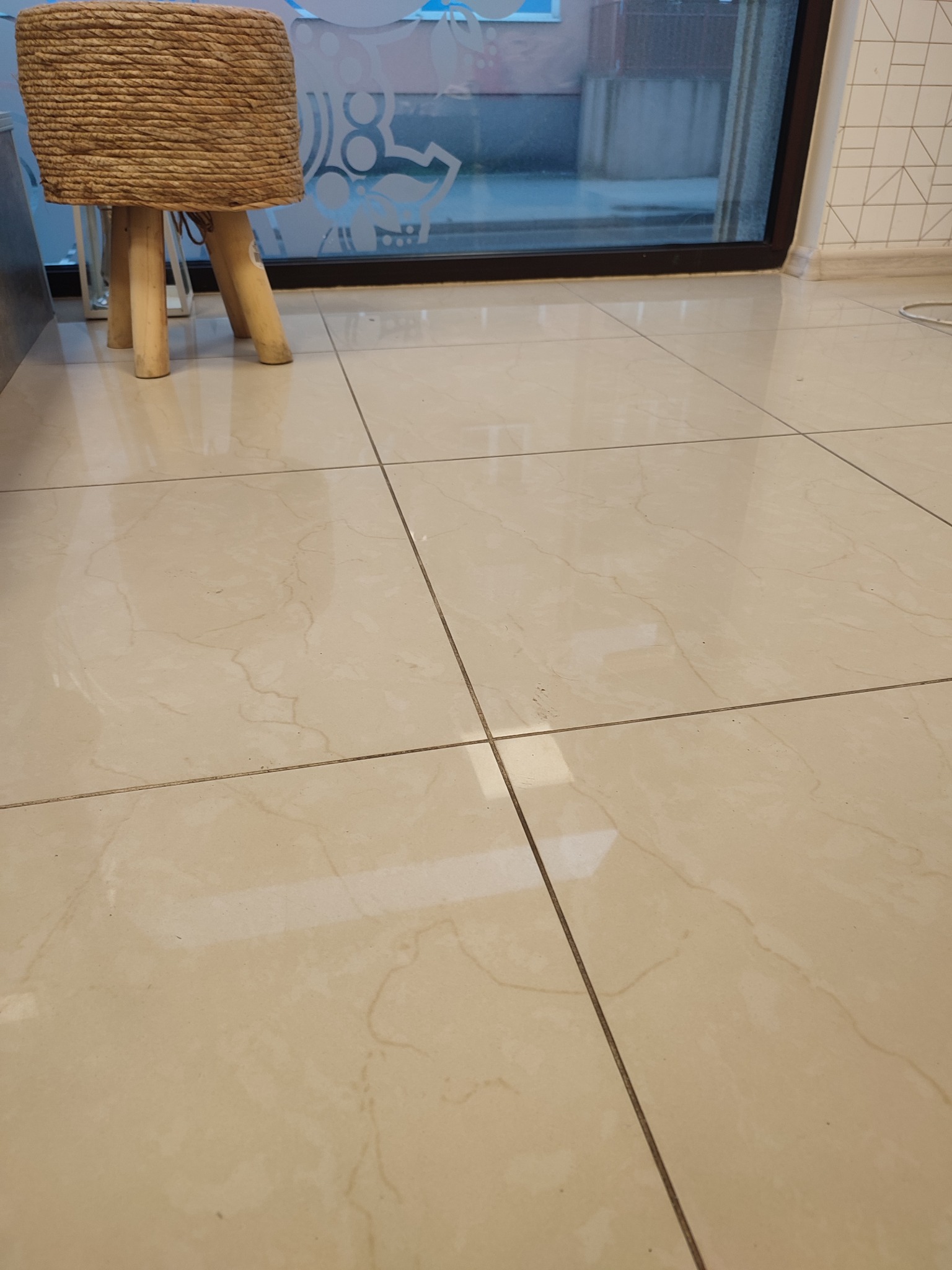 Beżowe płytki podłogowe z delikatnym wzorem marmuru, widoczne spomiędzy nóg plecionego taboretu z juty, z oknem w tle.