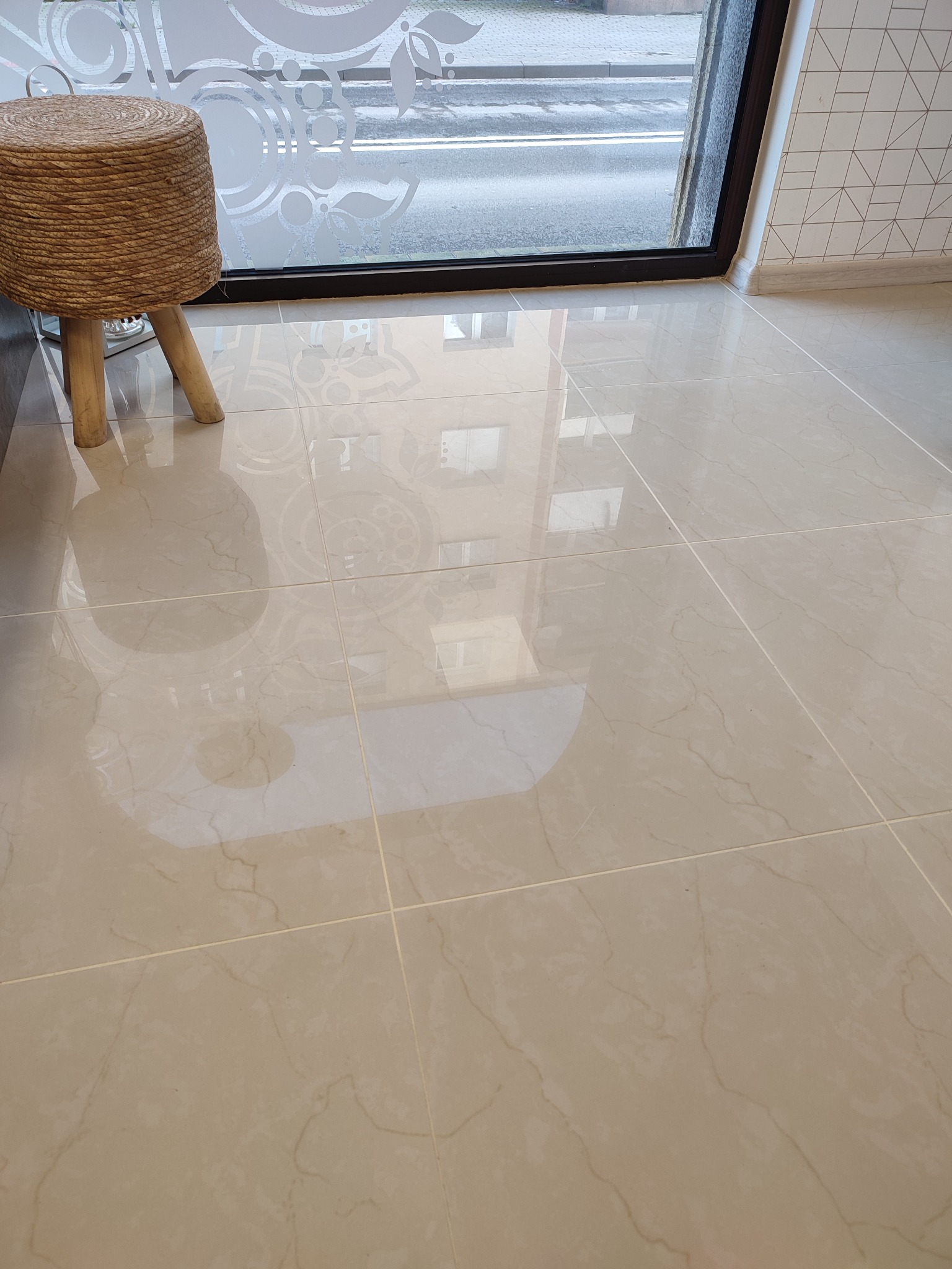 Beżowe płytki podłogowe z delikatnym marmurkowym wzorem, odbijające światło z okna, obok pleciony taboret na drewnianych nogach i fragment ściany z geometrycznym wzorem.