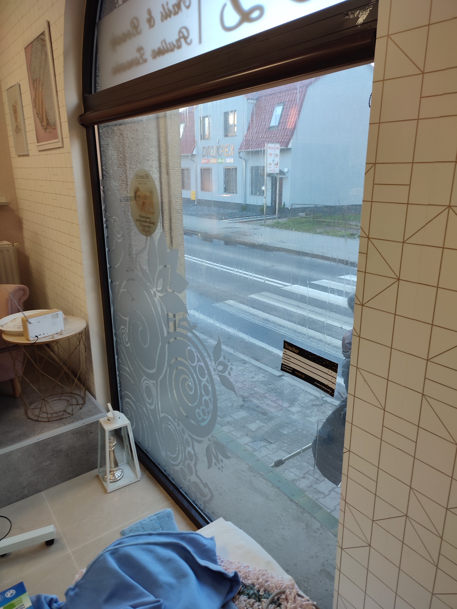 Wnętrze salonu kosmetycznego widziane przez okno z dekoracyjną folią, na pierwszym planie stolik z radiem i fragment niebieskiego ręcznika, za oknem ulica z przejściem dla pieszych i budynki.