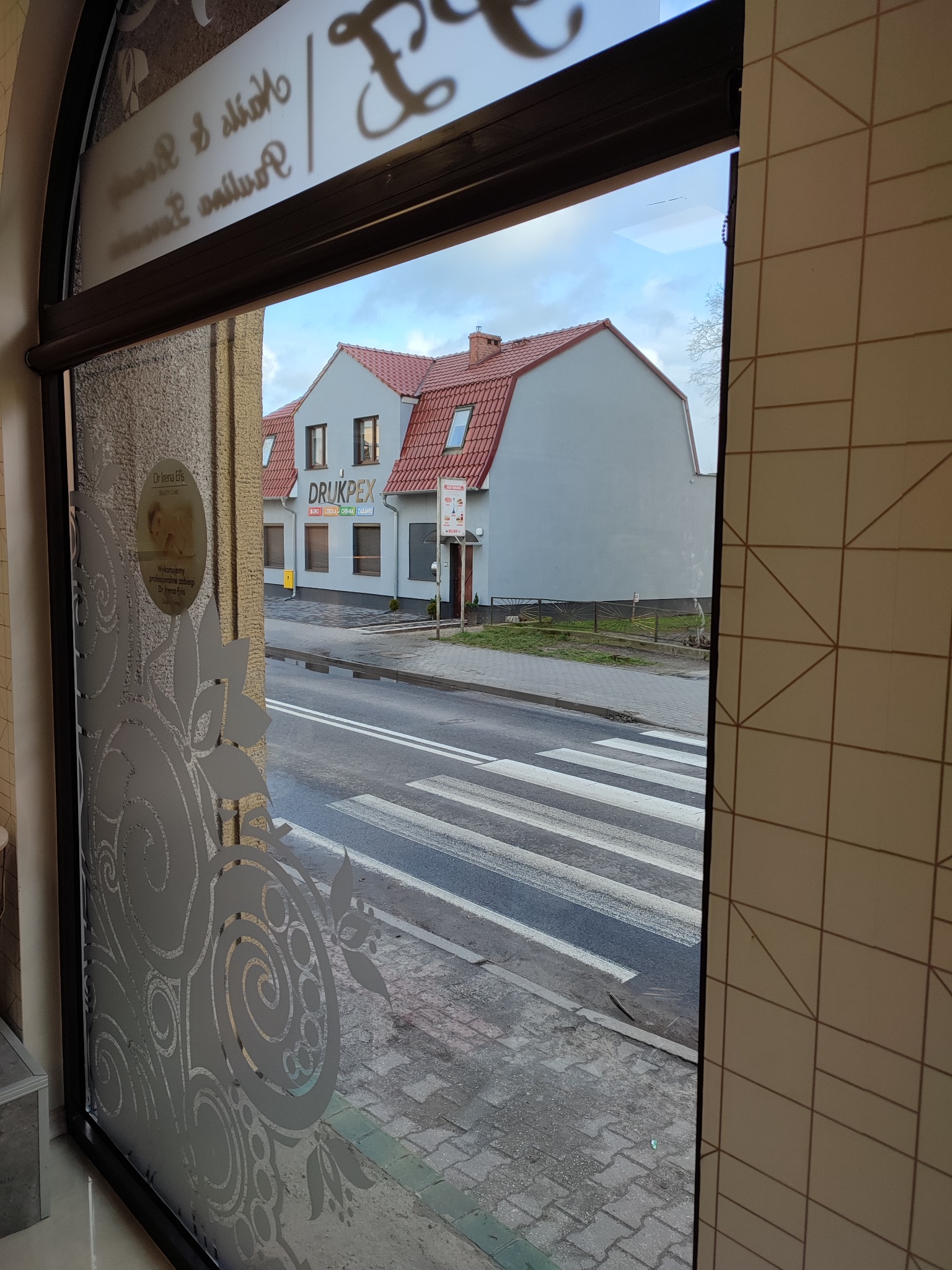 Widok z wnętrza budynku przez okno z dekoracyjną folią, na zewnątrz budynek z szyldem 'DRUKPEX' oraz przejście dla pieszych.