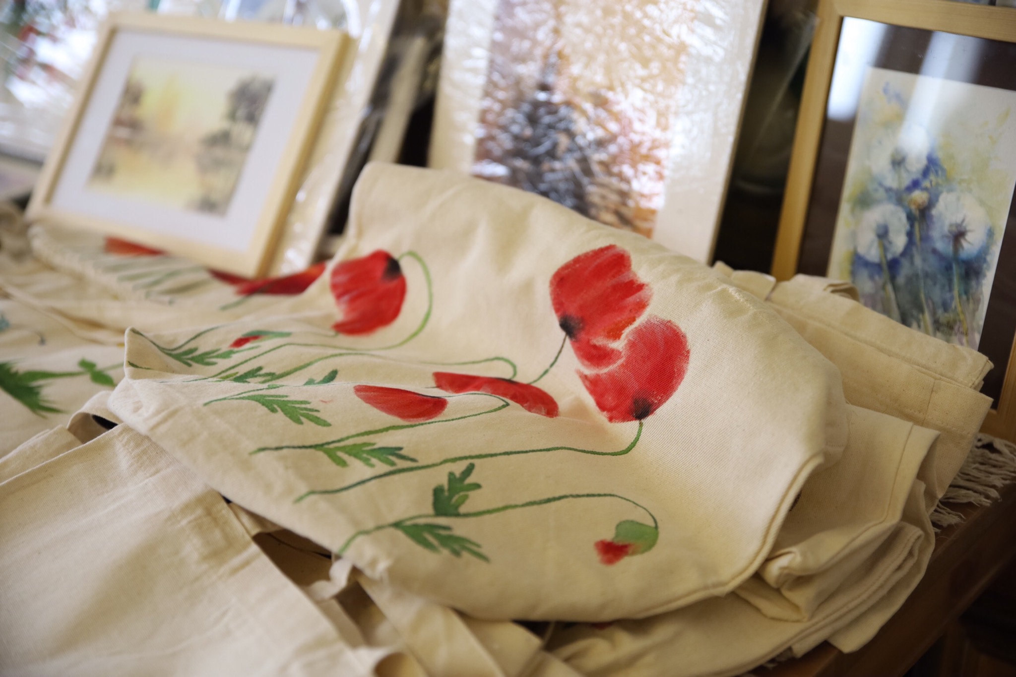 Beżowa torba bawełniana z ręcznie malowanymi czerwonymi makami i zielonymi łodygami, ułożona na drewnianym stole w Nowym Sączu, w tle ramki z obrazami.