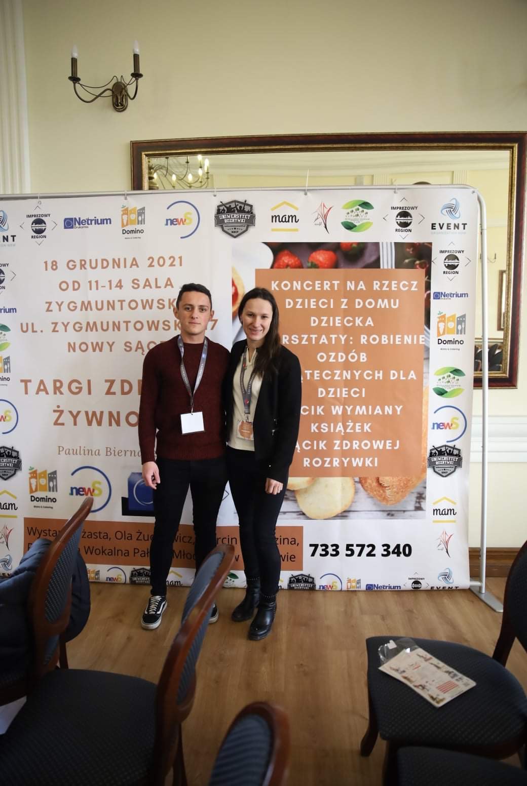 Dwie osoby pozują na tle ścianki reklamowej z informacjami o targach zdrowej żywności i wydarzeniach dla dzieci, w tym koncercie i warsztatach robienia ozdób.