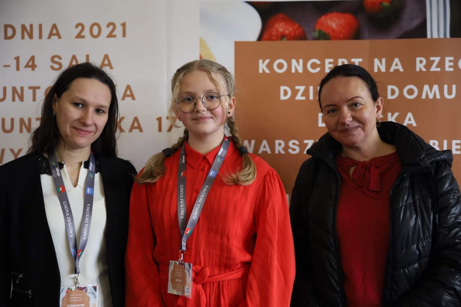 Trzy osoby pozują na tle plakatu z napisem 'Koncert na rzecz Dziecięcego Domu'. Osoby mają identyfikatory 'Gmina Iwkowa'.