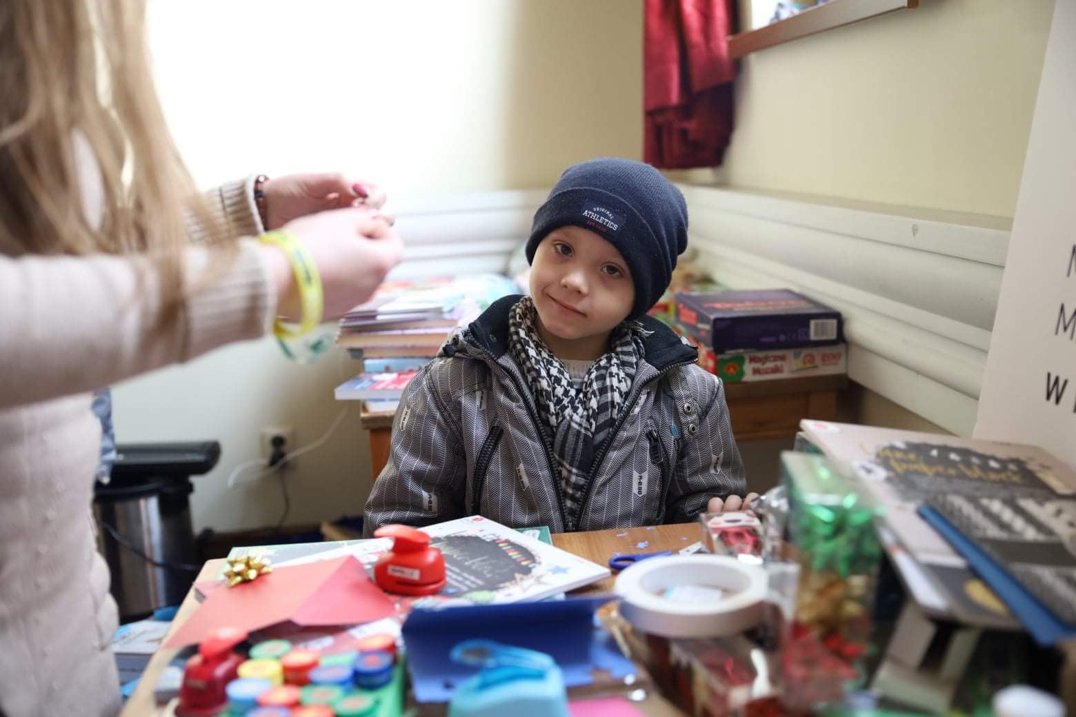 Chłopiec w czapce i kurtce siedzi przy stole pełnym materiałów do rękodzieła, w tle osoba przygotowuje ozdoby, tworząc świąteczną atmosferę.