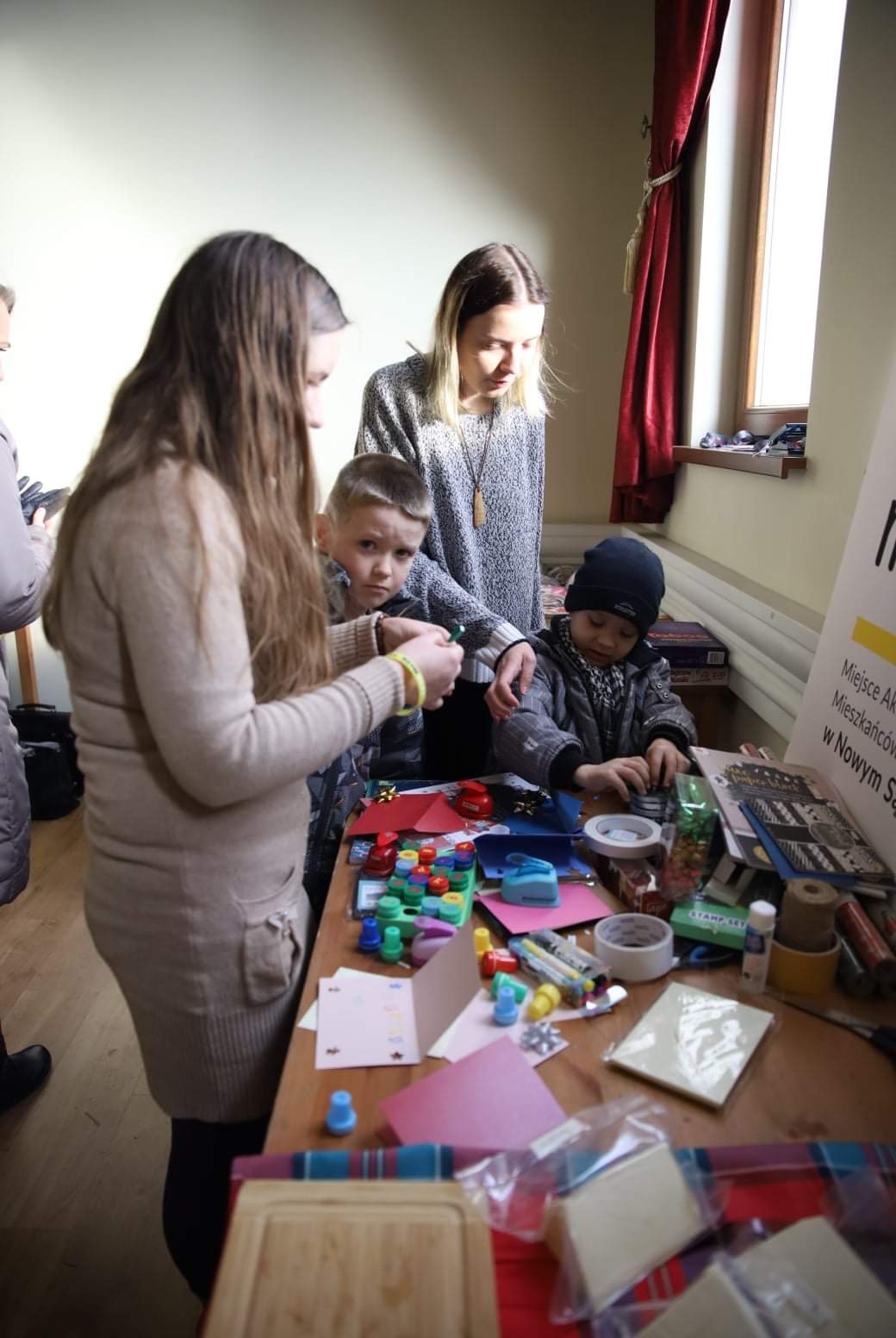 Dzieci z opiekunami tworzą kartki świąteczne przy stole zastawionym materiałami plastycznymi: kolorowe papiery, stempelki, wstążki, klej, nożyczki.