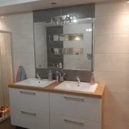 Nowoczesna łazienka z dwiema umywalkami nablatowymi na białych szafkach z drewnianym blatem, duże lustro z oświetleniem i półkami w tle, płytki w odcieniach szarości i beżu.