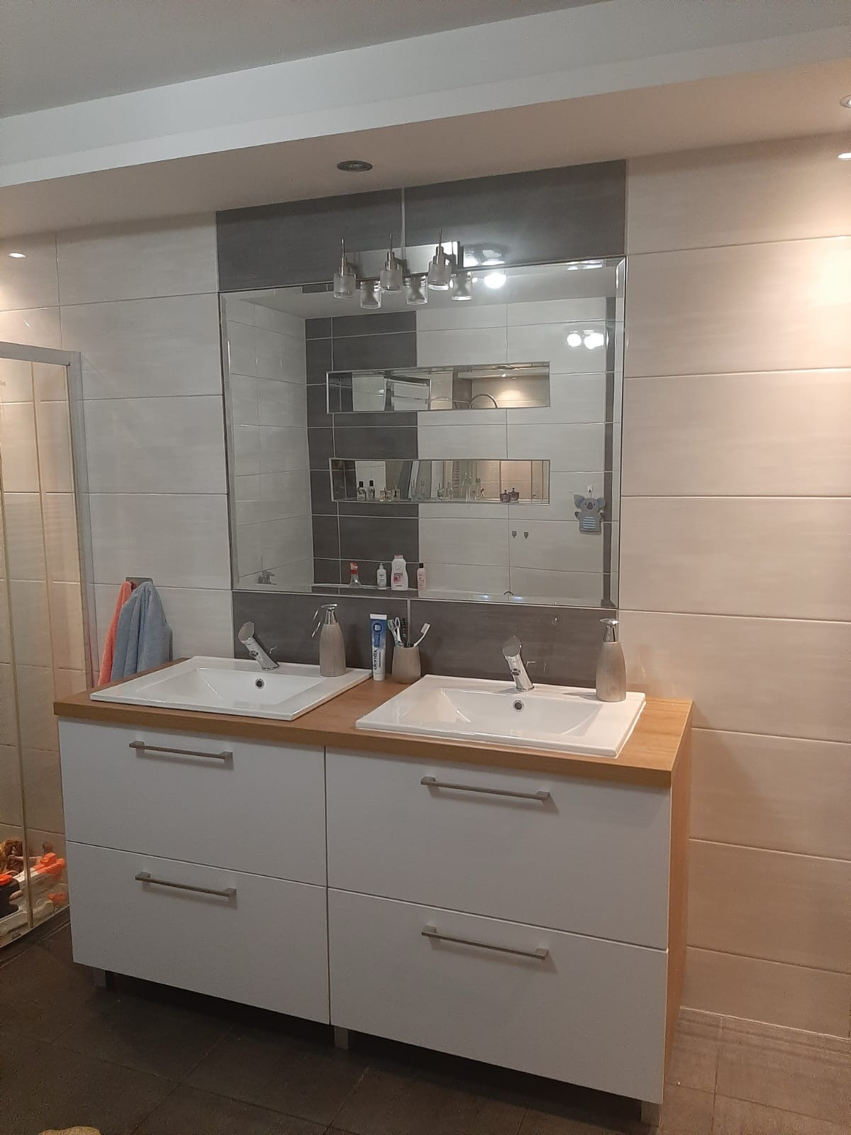 Nowoczesna łazienka z dwiema białymi szafkami pod umywalki z drewnianym blatem, duże lustro z oświetleniem i półkami w tle, płytki w odcieniach bieli i szarości.