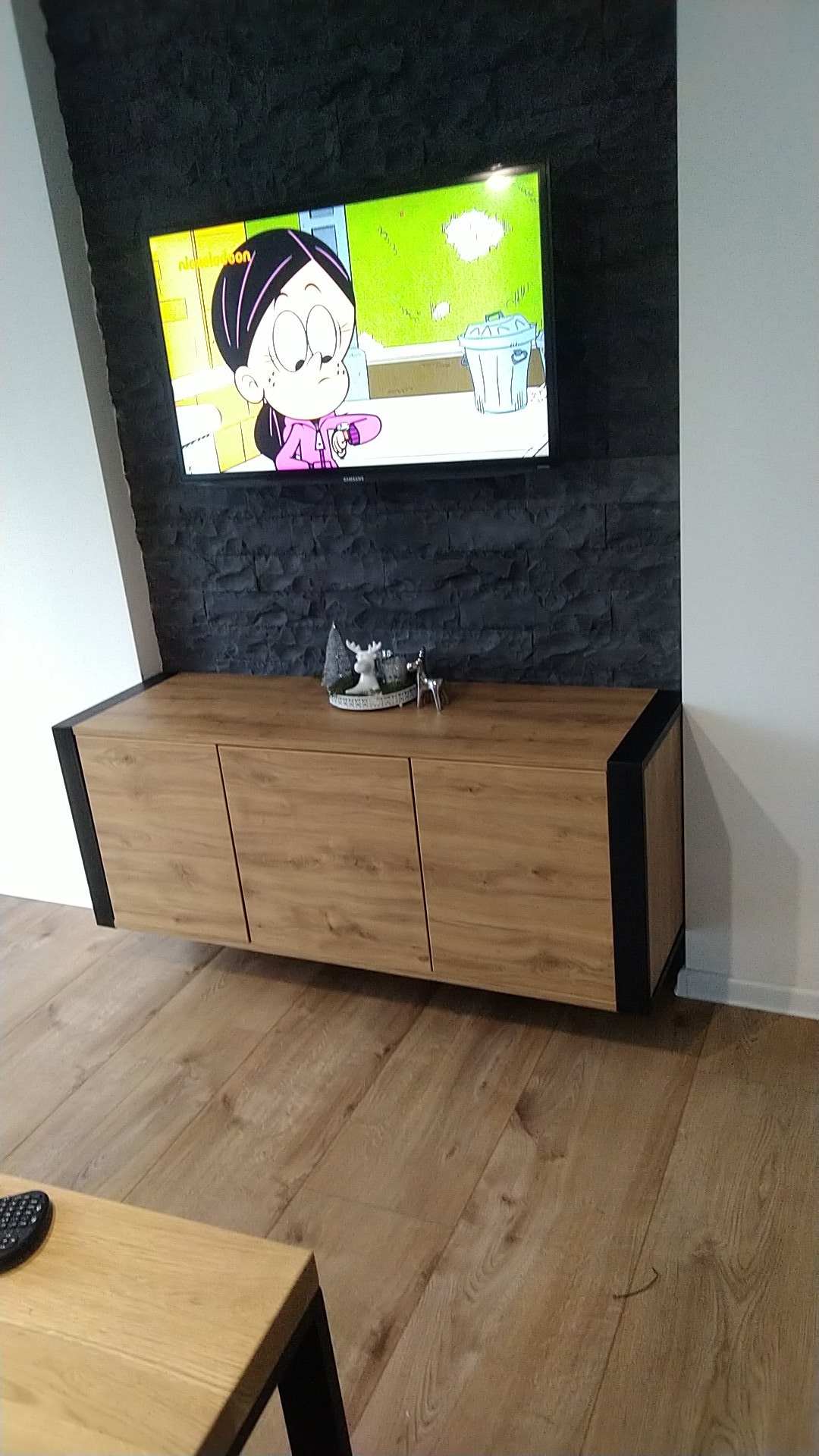 Nowoczesna szafka RTV z drewnianymi frontami i czarnymi bokami, zawieszona na ścianie z dekoracyjną okładziną kamienną, nad nią telewizor z włączoną kreskówką.