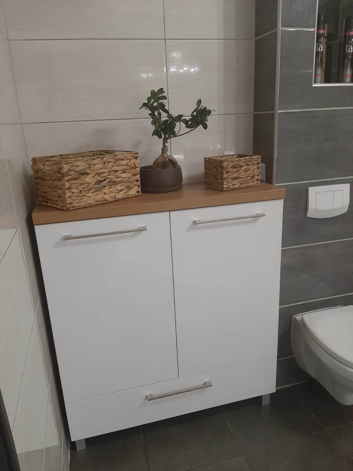 Biała szafka łazienkowa pod umywalkę z drewnianym blatem, na którym stoją dwa wiklinowe koszyki i małe drzewko bonsai w brązowej doniczce. Widoczny fragment toalety i szare płytki na ścianie.