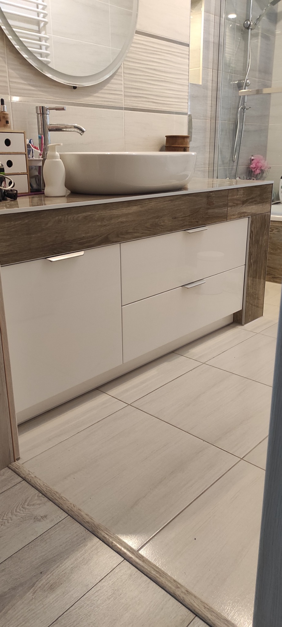 Nowoczesna szafka łazienkowa z białymi frontami i blatem imitującym drewno, okrągła umywalka nablatowa, widoczna kabina prysznicowa w tle.