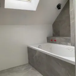 Nowoczesna łazienka w trakcie remontu z białą wanną zabezpieczoną folią, szarą glazurą na ścianach i podłodze, oraz oknem dachowym