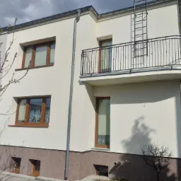 Elewacja budynku po renowacji z jasną farbą, widoczne okna z drewnianymi ramami, balkon z metalową balustradą i drabina ewakuacyjna, rynna spustowa.