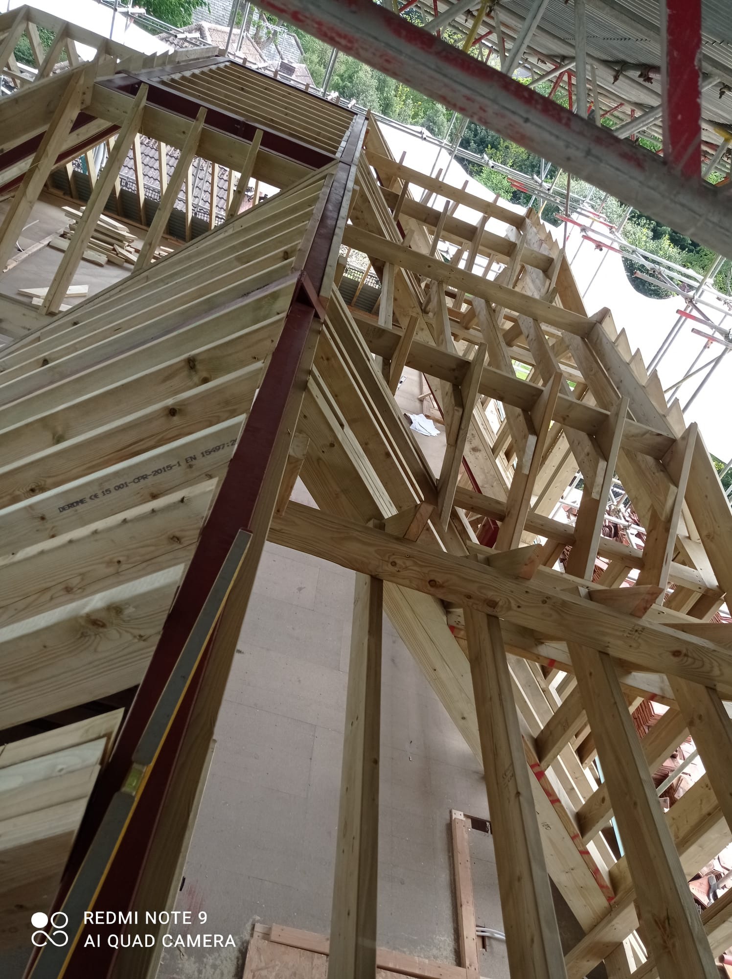 Konstrukcja drewniana więźby dachowej w trakcie montażu, widok od dołu, elementy stalowe wzmacniające konstrukcję, w tle widoczny fragment dachu sąsiedniego budynku.