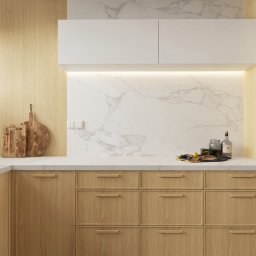 Studio Sto - Jasna kuchnia z drewnianymi szafkami, białym blatem i marmurowym backsplashiem, oświetlona listwą LED pod szafkami wiszącymi.
