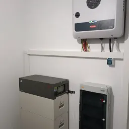 Wnętrze pomieszczenia z zainstalowanym systemem magazynowania energii: bateria BYD na podłodze, inwerter Fronius SYMO na ścianie, rozdzielnica elektryczna KENO obok, widoczne przewody i gniazdo...