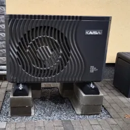 Zamontowana pompa ciepła KAISA na betonowych blokach, otoczona szarym żwirem i kostką brukową, z widocznym fragmentem elewacji budynku w tle.