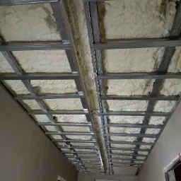 Metalowa konstrukcja sufitu podwieszanego z wypełnieniem pianką izolacyjną, widok z dołu, ściany w surowym tynku.